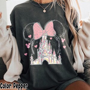 May include: A charcoal gray t-shirt featuring a graphic print of a castle silhouette topped with a large pink polka-dot bow and mouse ears. The design includes small pink hearts and firework outlines surrounding the central castle illustration.