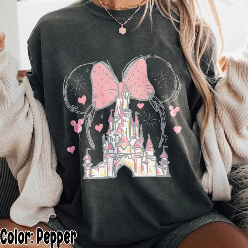 May include: A charcoal gray t-shirt featuring a graphic print of a castle silhouette topped with a large pink polka-dot bow and mouse ears. The design includes small pink hearts and firework outlines surrounding the central castle illustration.
