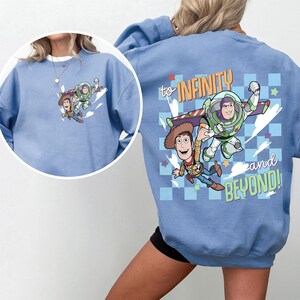 May include: Light blue sweatshirt featuring a graphic of Woody and Buzz Lightyear with the text "to INFINITY and BEYOND!". The design includes stars and a checkered pattern. The front of the sweatshirt also has a smaller version of the graphic.