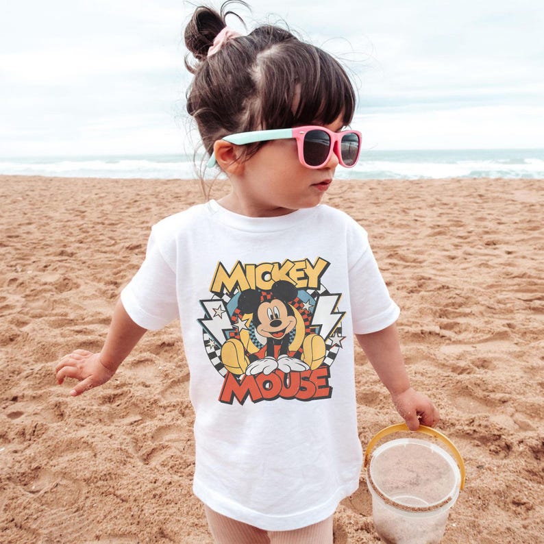 May include: A young child wearing a white t-shirt with a Mickey Mouse graphic, pink sunglasses, and a small bucket on a sandy beach. The shirt features the words "Mickey Mouse" in a colorful design.