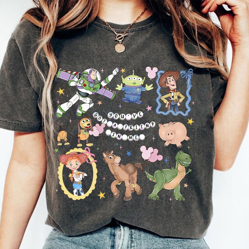 Pu&ograve; includere: T-shirt grigio scuro con personaggi di cartoni animati colorati. Il design include Buzz Lightyear, Woody, un alieno e altri personaggi, con la scritta "You've Got a Friend in Me".