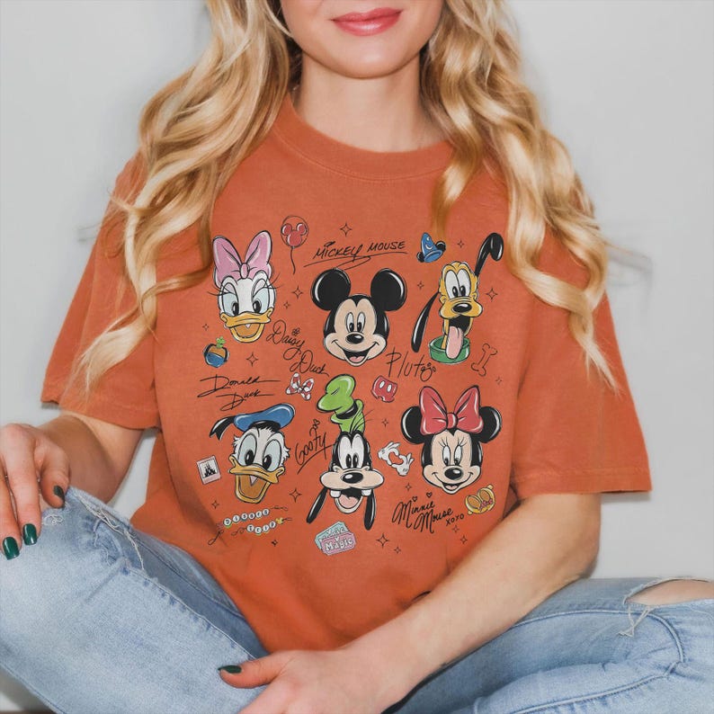 May include: Burnt orange t-shirt featuring a graphic of Mickey Mouse, Minnie Mouse, Donald Duck, Daisy Duck, Goofy, and Pluto. The design includes character signatures and other small graphics. The shirt is a relaxed fit.