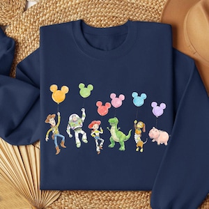 May include: Navy blue sweatshirt featuring a colorful illustration of cartoon characters holding Mickey Mouse-shaped balloons. The characters include a cowboy, a space ranger, a cowgirl, a green dinosaur, a dog, and a pig.