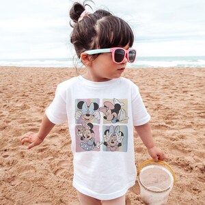 May include: White t-shirt featuring four squares with Minnie Mouse illustrations in pastel colors. The child is wearing pink sunglasses and is standing on a sandy beach. A small yellow bucket is in the child's hand.