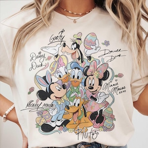 May include: Cream-colored t-shirt featuring a colorful graphic of Disney characters, including Mickey Mouse, Minnie Mouse, Donald Duck, and Goofy, with Easter-themed elements. The characters are surrounded by floral accents and their names are written in script.