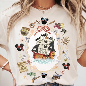 Könnte beinhalten: Cremefarbenes T-Shirt mit einem Piratenschiff-Design mit Mickey Mouse Ohren auf den Segeln. Das Design umfasst Piraten-Elemente wie eine Schatzkiste, einen Kompass und den Text "Pirates Life For Me."