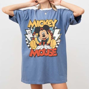 May include: Blue t-shirt featuring a large graphic of Mickey Mouse. The design includes the words "Mickey Mouse" in yellow and red, with Mickey in the center. The shirt has a white collar and short sleeves.