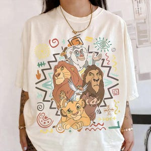 Puede incluir: Camiseta color crema con un gráfico colorido de personajes de El Rey León. El diseño incluye a Simba, Mufasa, Timón, Pumba y Rafiki, rodeados de patrones de estilo tribal en varios colores.