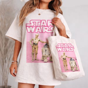 Könnte beinhalten: Cremefarbenes T-Shirt und Tragetasche mit einem rosa Star Wars-Design. Das Design zeigt das Star Wars-Logo und Bilder von C-3PO und R2-D2. Das T-Shirt hat kurze Ärmel.