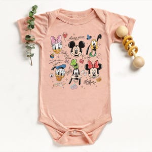 May include: A light pink baby onesie featuring a colorful graphic of Mickey Mouse and friends. The design includes Mickey, Minnie, Donald, Daisy, Goofy, and Pluto. The text "Mickey Mouse" is above the characters. A wooden rattle is on the right.