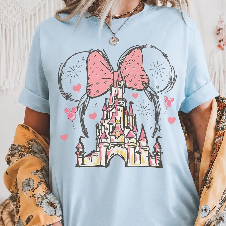 May include: A light blue short-sleeve t-shirt featuring a graphic print of a castle with a large pink polka-dot bow above it. The design includes black line-art outlines, small pink hearts, firework bursts, and Mickey Mouse-shaped balloons.