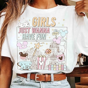 May include: White t-shirt with a colorful graphic design. The design includes the text "GIRLS JUST WANNA HAVE FUN" and illustrations of a castle, popcorn, a magic wand, and Mickey Mouse-themed items. The shirt has a crew neck.