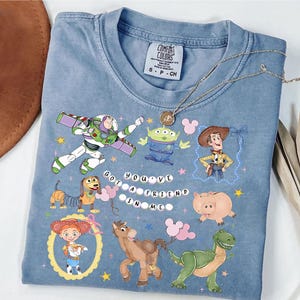 Pu&ograve; includere: T-shirt Comfort Colors azzurra con personaggi colorati di Toy Story. Il design presenta Buzz Lightyear, Woody e altri personaggi, con la scritta "You've Got a Friend in Me". Taglia S.