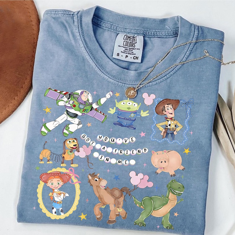Pu&ograve; includere: T-shirt Comfort Colors azzurra con personaggi colorati di Toy Story. Il design presenta Buzz Lightyear, Woody e altri personaggi, con la scritta "You've Got a Friend in Me". Taglia S.