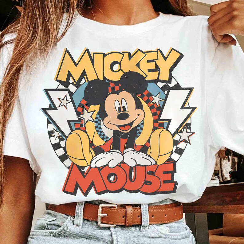 May include: White t-shirt featuring a colorful graphic of Mickey Mouse. The design includes the words "Mickey Mouse" in yellow and red, with a retro-style background of stars, lightning bolts, and a checkered pattern. A brown belt is visible.