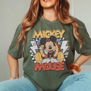 May include: Olive green t-shirt featuring a graphic of Mickey Mouse. The design includes the words "Mickey Mouse" in yellow and red, with a cartoon image of Mickey. The shirt has short sleeves and a crew neck.