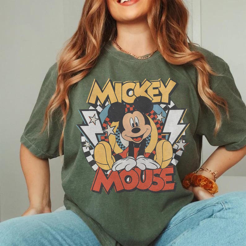 May include: Olive green t-shirt featuring a graphic of Mickey Mouse. The design includes the words "Mickey Mouse" in yellow and red, with a cartoon image of Mickey. The shirt has short sleeves and a crew neck.