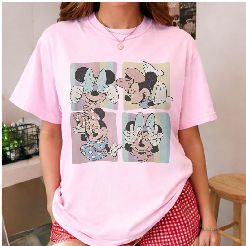 May include: A light pink t-shirt featuring a four-panel graphic of Minnie Mouse in various poses. Each panel has a pastel background. Minnie is wearing a bow and a dress in the bottom left panel. The shirt has short sleeves.