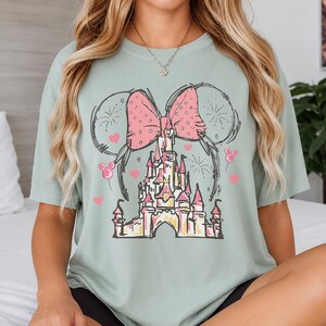 May include: A sage green short-sleeve t-shirt featuring a graphic print of a castle with a large pink polka-dot bow above it. The design includes black line-art outlines of mouse ears, small pink hearts, and fireworks surrounding the castle.