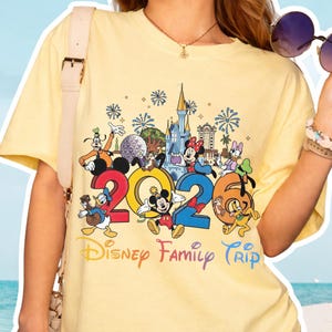 Könnte beinhalten: Ein hellgelbes T-Shirt mit einer farbenfrohen Grafik von Disney-Figuren und dem Text "2020 Disney Family Trip". Das Design umfasst Micky Maus, Minnie Maus, Donald Duck, Goofy und andere Charaktere sowie Feuerwerk und ein Schloss.