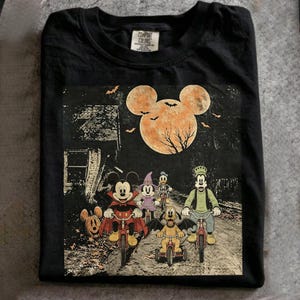 Pode incluir: Uma t-shirt preta com um gráfico do Mickey, Minnie, Pato Donald, Pateta e Pluto a andar de bicicleta num caminho à noite. Uma grande lua cor de laranja em forma de cabeça do Mickey, morcegos e uma casa estão visíveis no fundo.