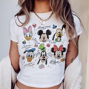 May include: Cream-colored t-shirt featuring cartoon character illustrations of Mickey Mouse, Minnie Mouse, Donald Duck, Daisy Duck, Goofy, and Pluto. The shirt has handwritten character names and decorative elements.