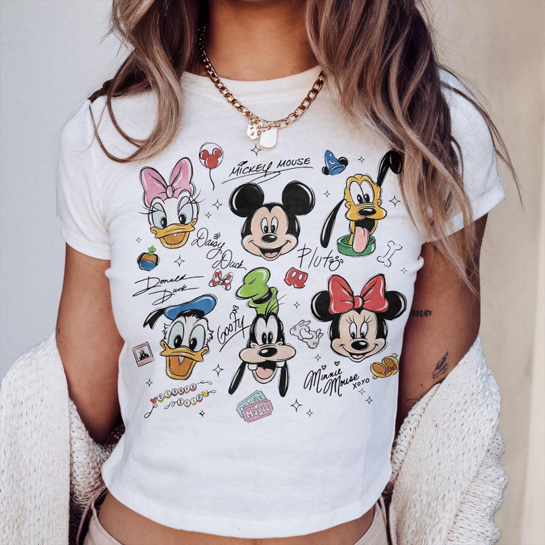 May include: Cream-colored t-shirt featuring cartoon character illustrations of Mickey Mouse, Minnie Mouse, Donald Duck, Daisy Duck, Goofy, and Pluto. The shirt has handwritten character names and decorative elements.