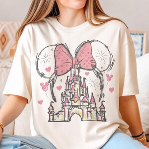 May include: A cream-colored short-sleeve t-shirt featuring a black line-art graphic of a castle topped with a large pink polka-dot bow and mouse ears. The design includes small pink hearts, firework bursts, and mouse-shaped balloon icons.
