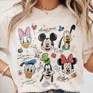 May include: Cream-colored t-shirt featuring cartoon illustrations of Mickey Mouse, Minnie Mouse, Donald Duck, Daisy Duck, Goofy, and Pluto. The characters' names are written in cursive script. The shirt has a crew neck.
