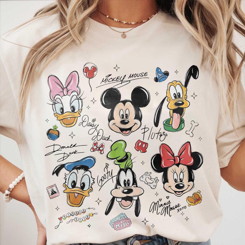 May include: Cream-colored t-shirt featuring cartoon illustrations of Mickey Mouse, Minnie Mouse, Donald Duck, Daisy Duck, Goofy, and Pluto. The characters' names are written in cursive script. The shirt has a crew neck.