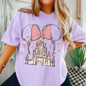 May include: A light purple t-shirt featuring a graphic print of a castle topped with a large polka-dot bow and mouse ears. The design includes line-art fireworks, small hearts, and balloon icons in shades of pink, white, and black.