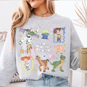 Pu&ograve; includere: Una felpa grigio chiaro con personaggi di cartoni animati colorati del film Toy Story. Il design include Buzz Lightyear, Woody e altri personaggi, con la scritta "You've Got a Friend in Me".