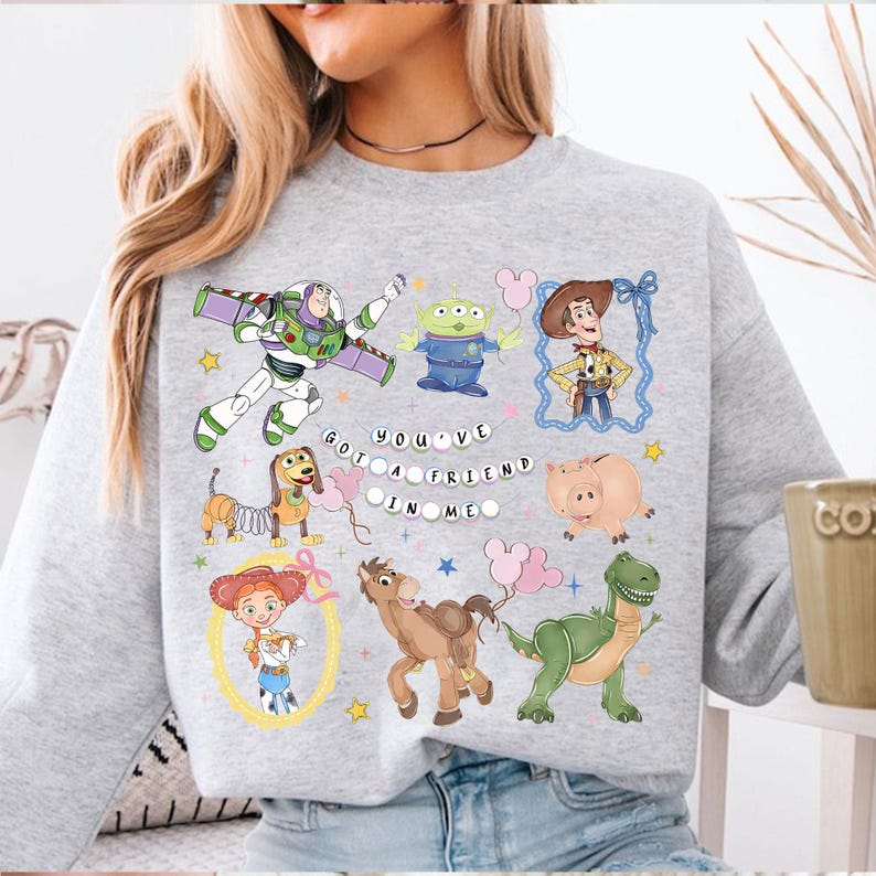 Pu&ograve; includere: Una felpa grigio chiaro con personaggi di cartoni animati colorati del film Toy Story. Il design include Buzz Lightyear, Woody e altri personaggi, con la scritta "You've Got a Friend in Me".