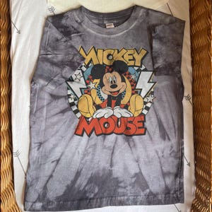 May include: A gray tie-dye t-shirt featuring a graphic of Mickey Mouse. The design includes the words "Mickey Mouse" in yellow and red, with Mickey in a seated pose. The shirt has a crew neck and short sleeves.