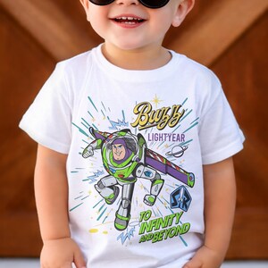 May include: A white short-sleeved t-shirt featuring a graphic of Buzz Lightyear in his green, white, and purple space suit. The design includes the text Buzz Lightyear and To Infinity and Beyond in yellow and green, with blue starburst accents.