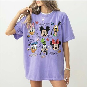 May include: A lilac-colored t-shirt featuring a graphic of Mickey Mouse and friends. The design includes images of Mickey, Minnie, Donald, Daisy, Goofy, and Pluto, along with their signatures. The shirt has short sleeves and a crew neck.
