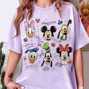 May include: A light purple t-shirt featuring various Disney cartoon characters, including Mickey Mouse, Minnie Mouse, Donald Duck, Daisy Duck, Goofy, and Pluto. The shirt has a crew neck and short sleeves. The characters are in a cartoon style.