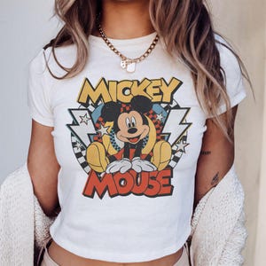 May include: White t-shirt featuring a vintage-style graphic of Mickey Mouse. The design includes the words "Mickey Mouse" in bold lettering, with Mickey in a seated pose. The background has lightning bolts, stars, and a checkered pattern.