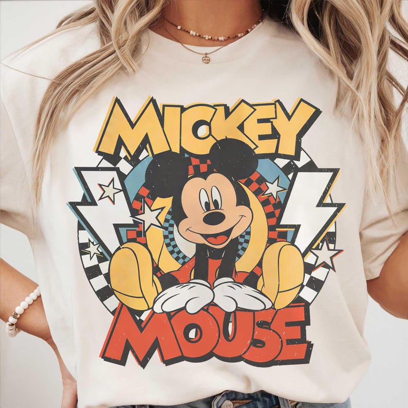 May include: Cream-colored t-shirt featuring a retro graphic of Mickey Mouse. The design includes the words "Mickey Mouse" in bold, colorful lettering, with Mickey in the center. The graphic has a vintage, cartoon style.