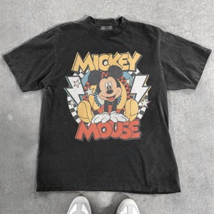 May include: A black t-shirt featuring a graphic of Mickey Mouse. The design includes the words "Mickey Mouse" in red and yellow, with Mickey in a seated pose. The shirt has a vintage, worn look.