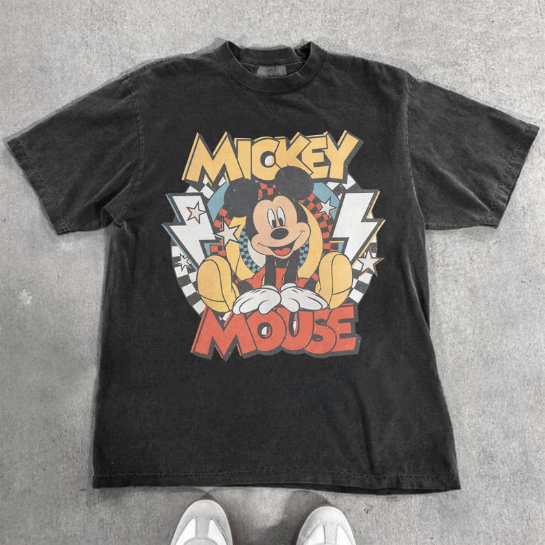 May include: A black t-shirt featuring a graphic of Mickey Mouse. The design includes the words "Mickey Mouse" in red and yellow, with Mickey in a seated pose. The shirt has a vintage, worn look.
