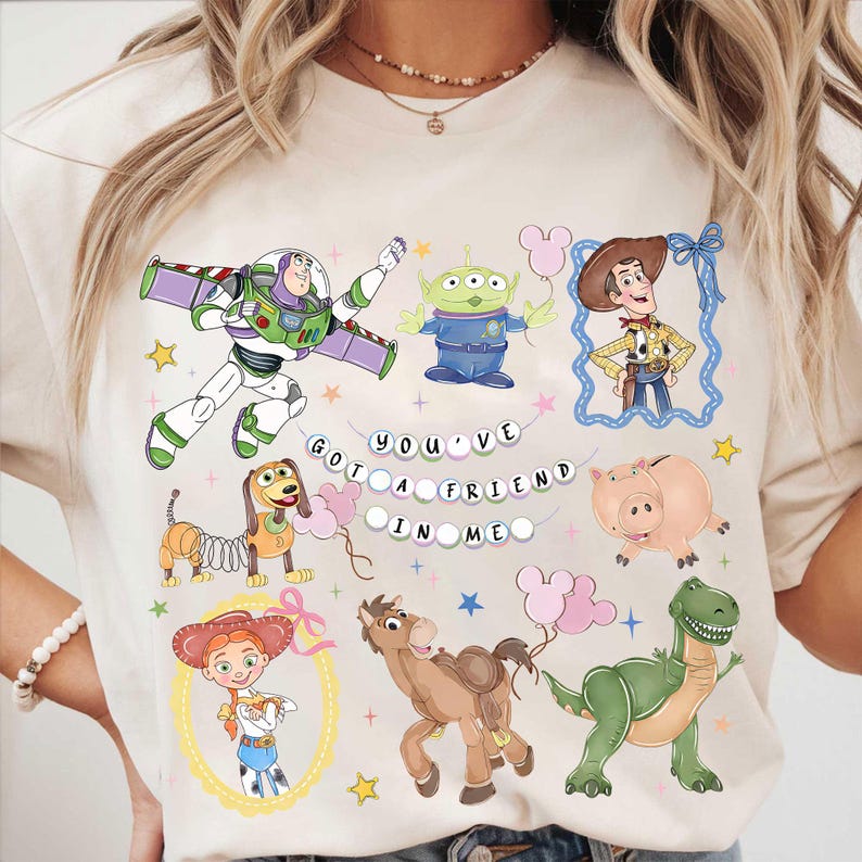 Pu&ograve; includere: T-shirt color crema con personaggi di cartoni animati colorati di un popolare film d'animazione. Il design include Buzz Lightyear, Woody, un alieno, Jessie, un maiale, un dinosauro, un cavallo e un bassotto. Il testo recita "You've Got a Friend in Me."