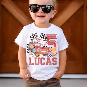 May include: White t-shirt with a red race car graphic, chequered flags, the number 5, and the name "LUCAS" in red. A young child wearing black sunglasses is wearing the shirt.