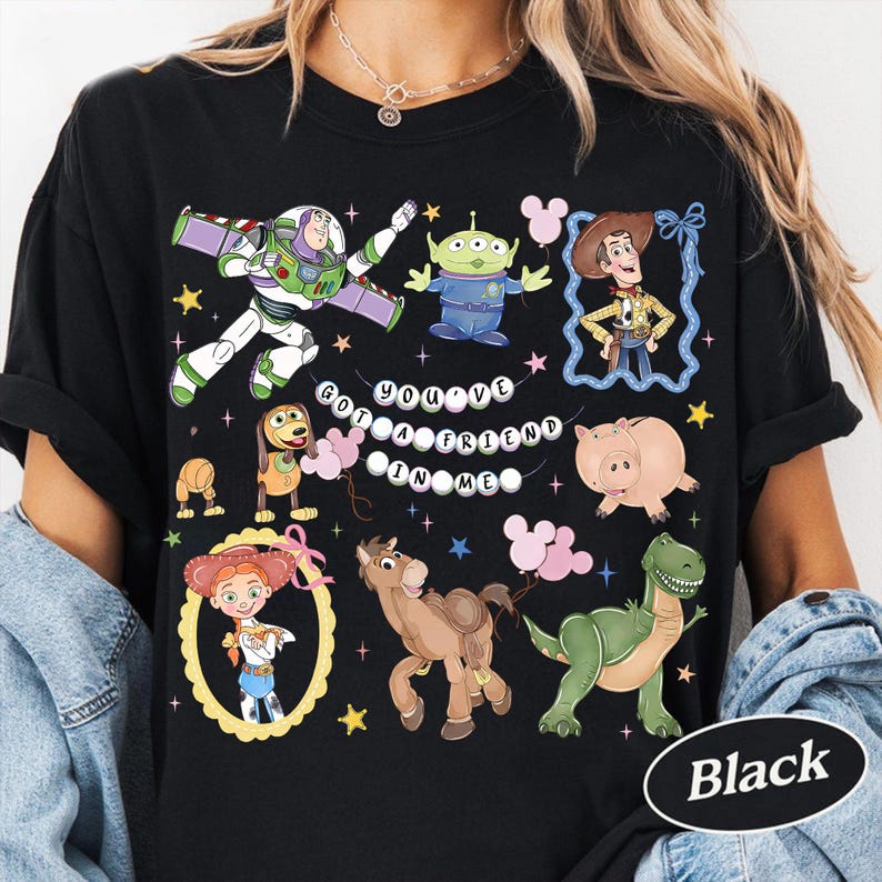 Pu&ograve; includere: T-shirt nera con personaggi di cartoni animati colorati e la scritta "YOU'VE GOT A FRIEND IN ME." Il design include Buzz Lightyear, Woody e altri personaggi. Un ovale nero con la parola "Black" &egrave; in basso a destra.