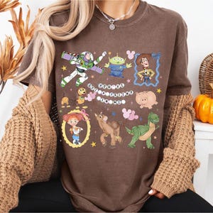 Pu&ograve; includere: T-shirt marrone con personaggi dei cartoni animati tra cui Buzz Lightyear, Woody e i piccoli omini verdi. Il testo "YOU'VE GOT A FRIEND IN ME" &egrave; visualizzato. La maglietta ha un girocollo e maniche corte.
