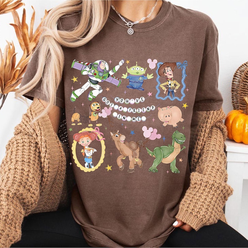 Pu&ograve; includere: T-shirt marrone con personaggi dei cartoni animati tra cui Buzz Lightyear, Woody e i piccoli omini verdi. Il testo "YOU'VE GOT A FRIEND IN ME" &egrave; visualizzato. La maglietta ha un girocollo e maniche corte.