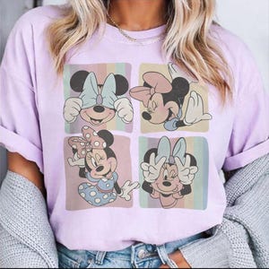 May include: A light purple t-shirt featuring a four-panel graphic of Minnie Mouse in various poses. Each panel has a different background color with pastel stripes. Minnie is wearing bows and dresses in the graphic.