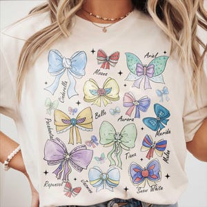 May include: A cream-coloured t-shirt displaying a variety of colourful bow illustrations. Each bow is a different colour and style, with names such as Cinderella, Ariel, and Belle written below. The design includes stars and a whimsical aesthetic.