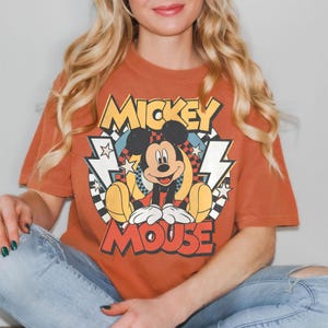 May include: Burnt orange t-shirt featuring a vintage Mickey Mouse graphic. The design includes the words "Mickey Mouse" in yellow and red, with Mickey Mouse sitting in the center. The shirt is paired with blue jeans.