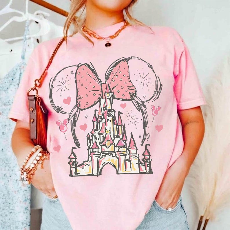 May include: A light pink short-sleeve t-shirt featuring a graphic print of a castle silhouette topped with a large polka-dot bow and mouse ears. The design includes small heart shapes and firework bursts in black and pink ink outlines.